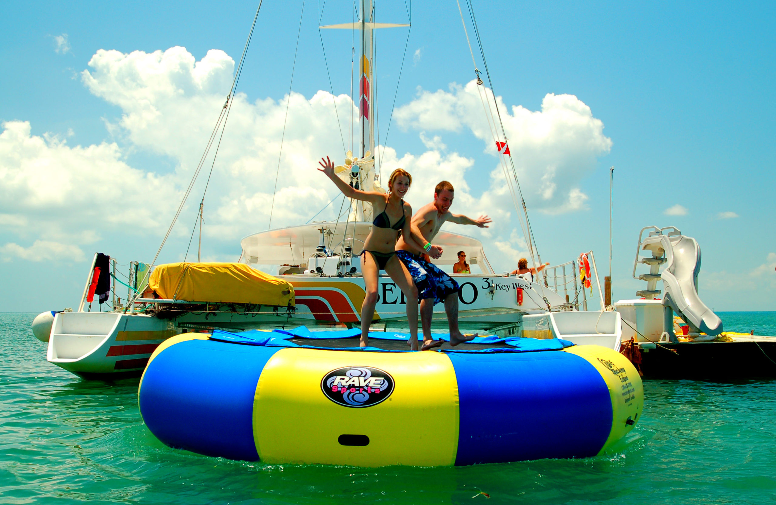 A catamaran with people playing on waterways including a trampoline, slide and kayaks